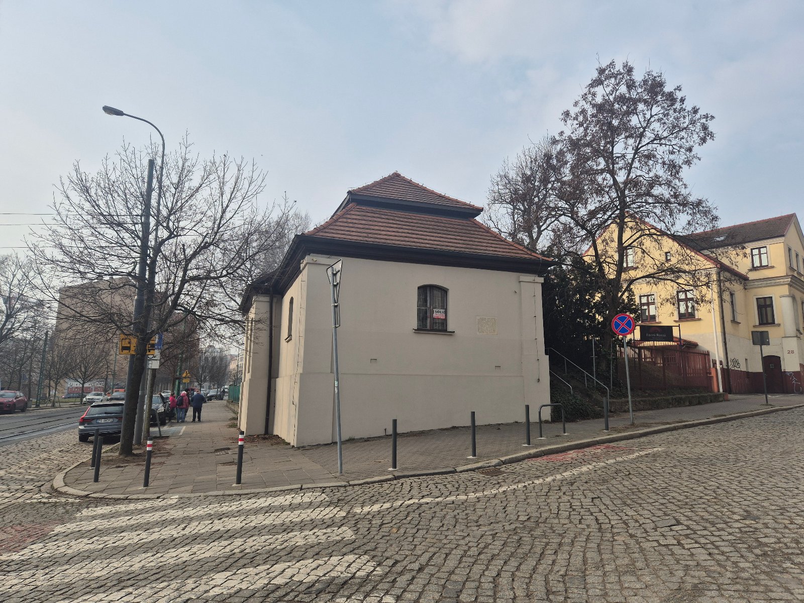 Zdjęcie przedstawia budynek. Obok znajduje się inny budynek oraz ulica przy której zaparkowane są samochody.