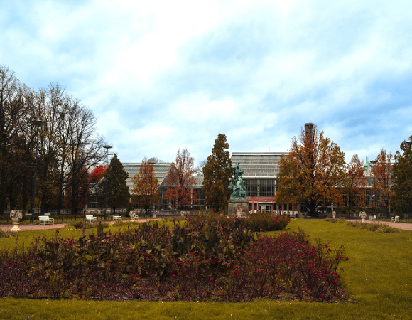 Zdjęcie przedstawia budynek palmiarni i otaczającą ją zieleń oraz stojacy obok pomnik.