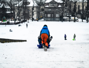 Zdjęcie przedstawia chłopca pchajacego kolegę na sankach z górki. W oddali widać inne dzieci.