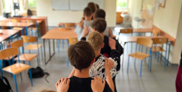 Zdjęcie przedstawia chłopców stojących w rzędzie, trzymając się za barki, tyłem do aparatu. Zdjęcie zrobiono w klasie lekcyjnej. W tle widać ławki i krzesła.