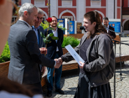 Zdjęcie przedstawia czterech mężczyzn. Jeden z nich ściska rękę dziewczynie i wręcza jej różę. Zdjęcie zrobiono przed urzędem. W tle widać budynek i zieleń.