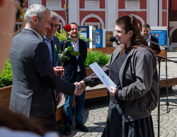 Zdjęcie przedstawia czterech mężczyzn. Jeden z nich ściska rękę dziewczynie i wręcza jej różę. Zdjęcie zrobiono przed urzędem. W tle widać budynek i zieleń.