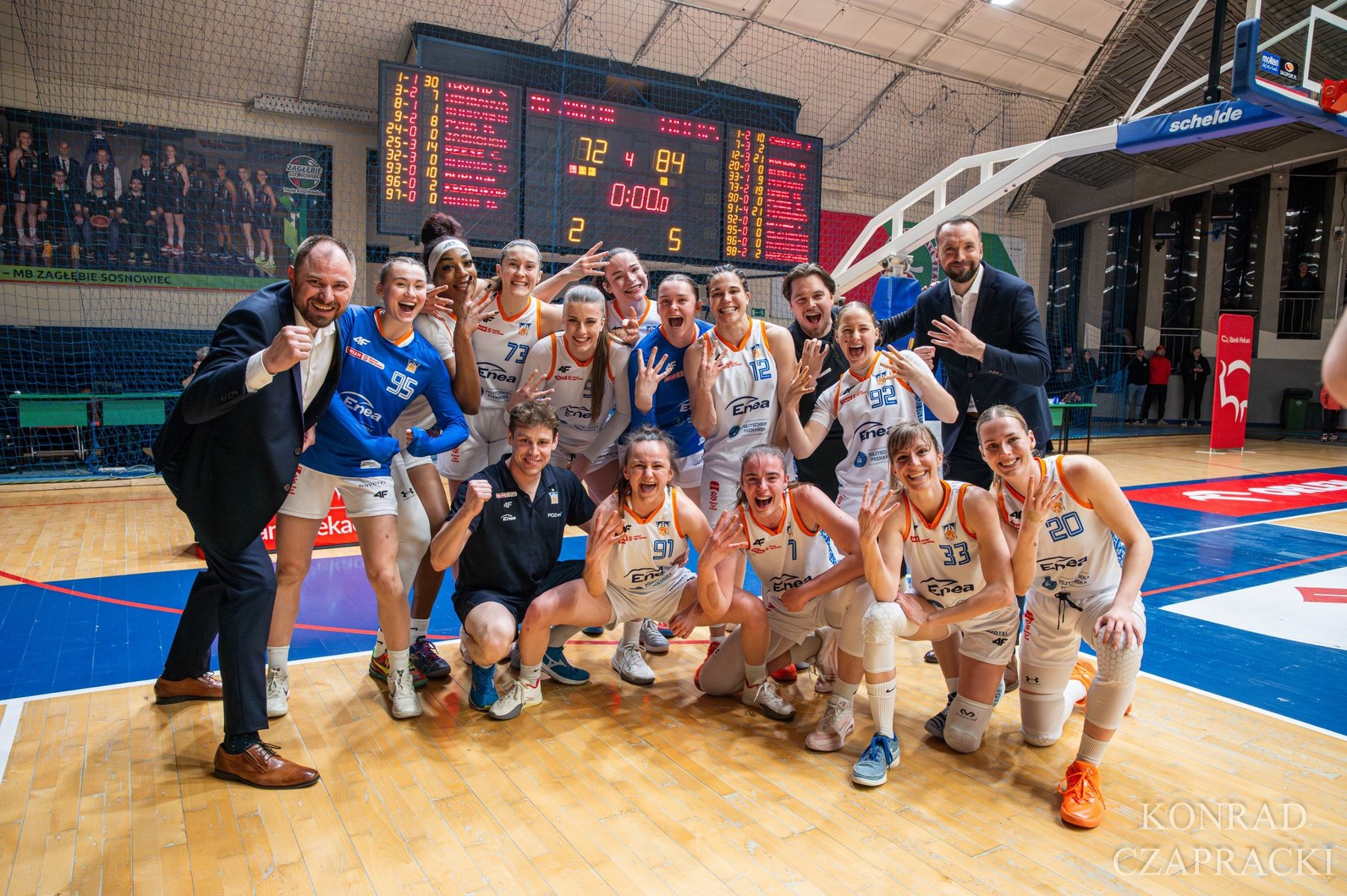 Zdjęcie przedstawia drużynę koszykarską Enea świętującą zwycięstwo na parkiecie hali sportowej. Zawodniczki i sztab szkoleniowy pozują do wspólnego zdjęcia w radosnych pozach, wykonując gesty zwycięstwa. W tle widoczna jest duża tablica wyników, która wskazuje końcowy rezultat meczu 72:84 przy czasie 0:00.