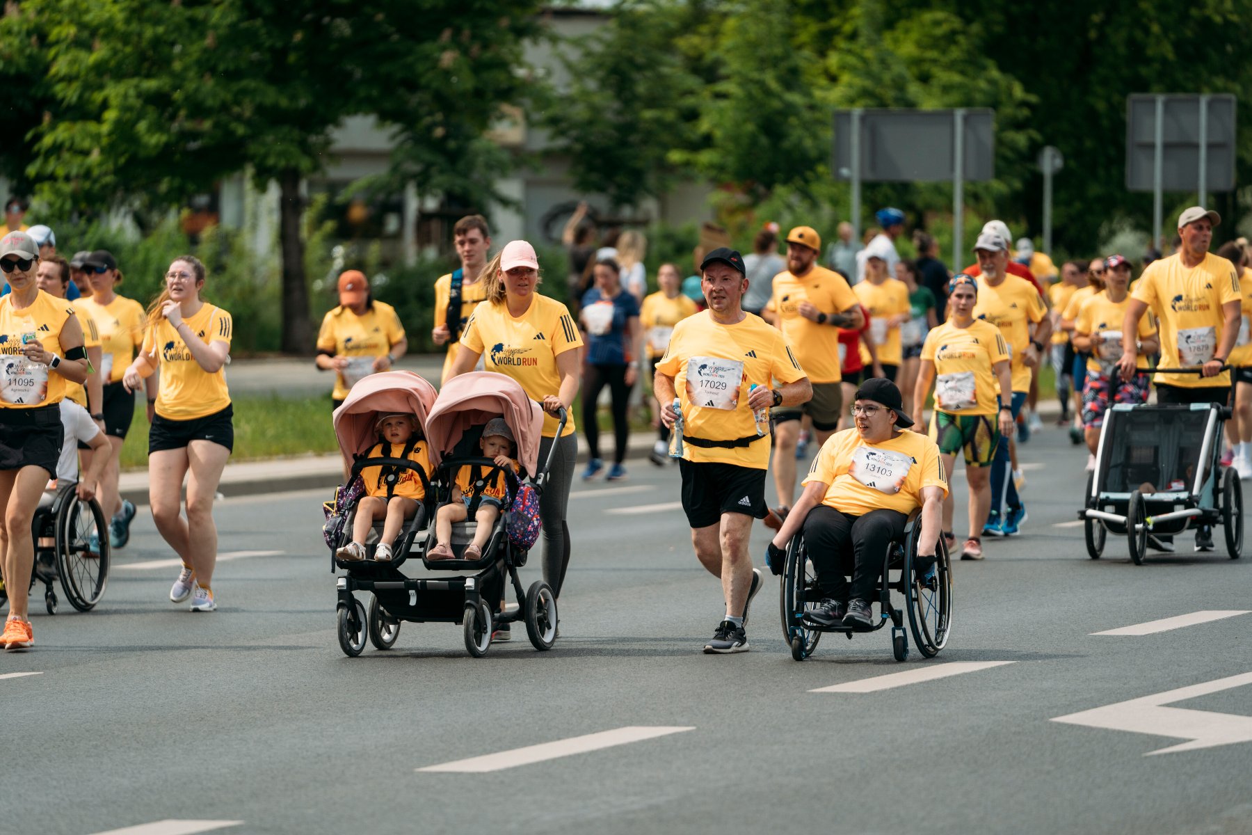 Zdjęcie przedstawia dużą grupę uczestników biegu charytatywnego Wings for Life World Run, poruszających się po szerokiej asfaltowej ulicy. Większość osób ma na sobie charakterystyczne żółte koszulki techniczne z logo biegu oraz numery startowe przypięte na piersiach. W centrum kadru widać kobietę pchającą podwójny wózek dziecięcy z dwójką dzieci, a obok niej biegnących mężczyzn oraz osobę poruszającą się na wózku inwalidzkim.