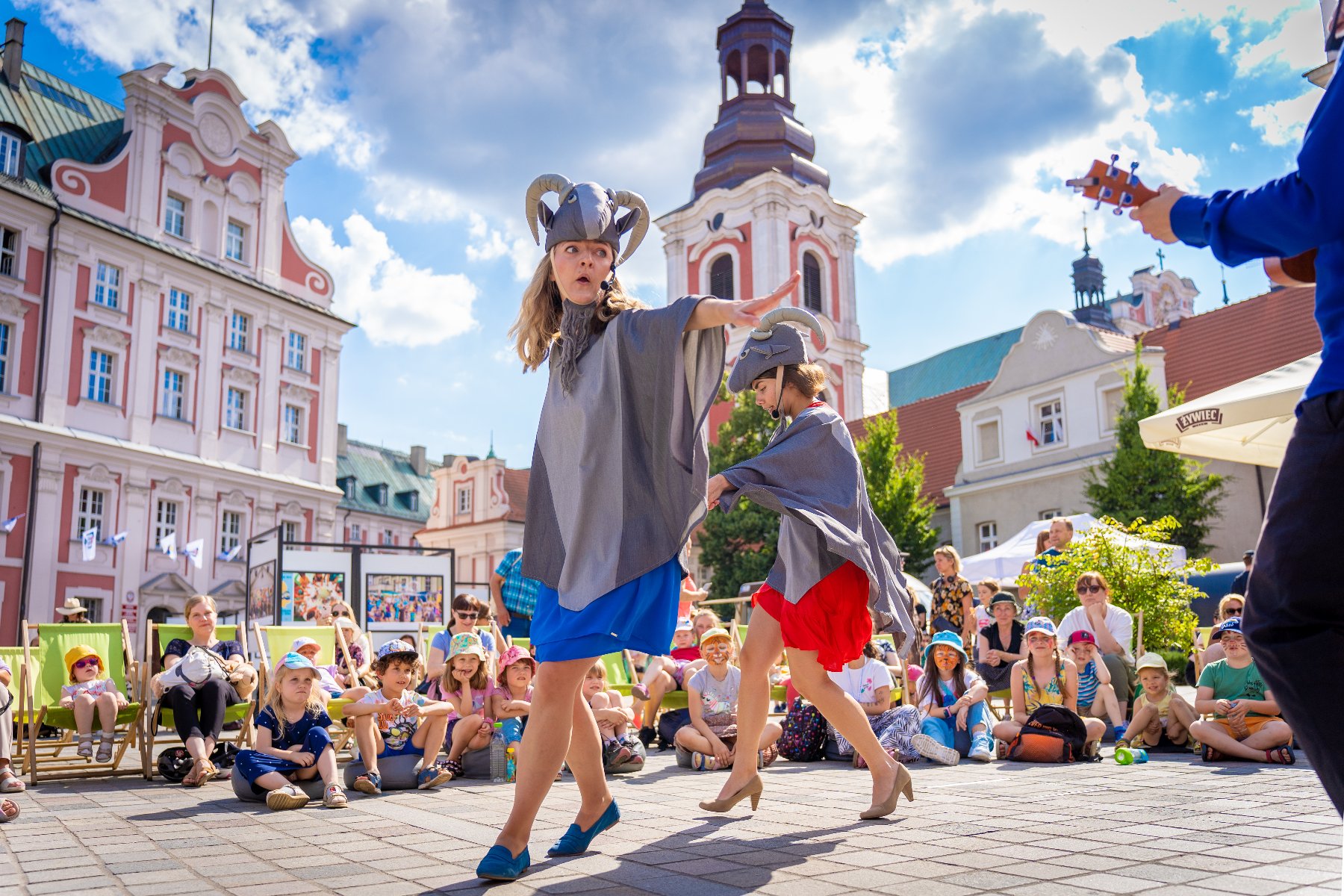Zdjęcie przedstawia dwie kobiety przebrane za koziołki oraz tłum patrzących na nie dzieci. W tle widac budynek urzędu miasta.