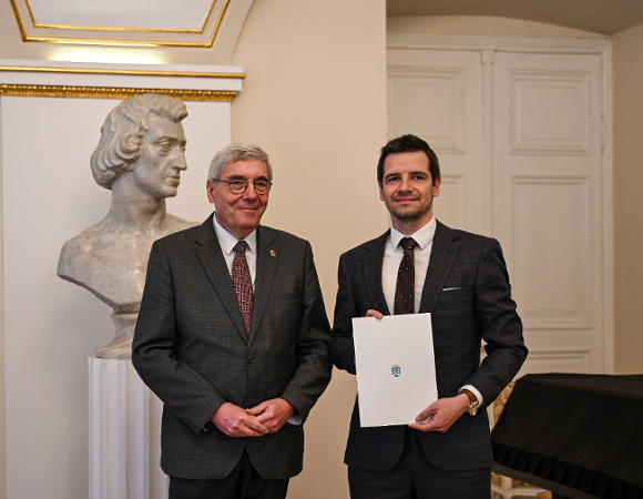 Zdjęcie przedstawia dwóch mężczyzn - młodszego i starszego - pozyjących do zdjęcia. Młodszy z nich trzyma w ręku teczkę.