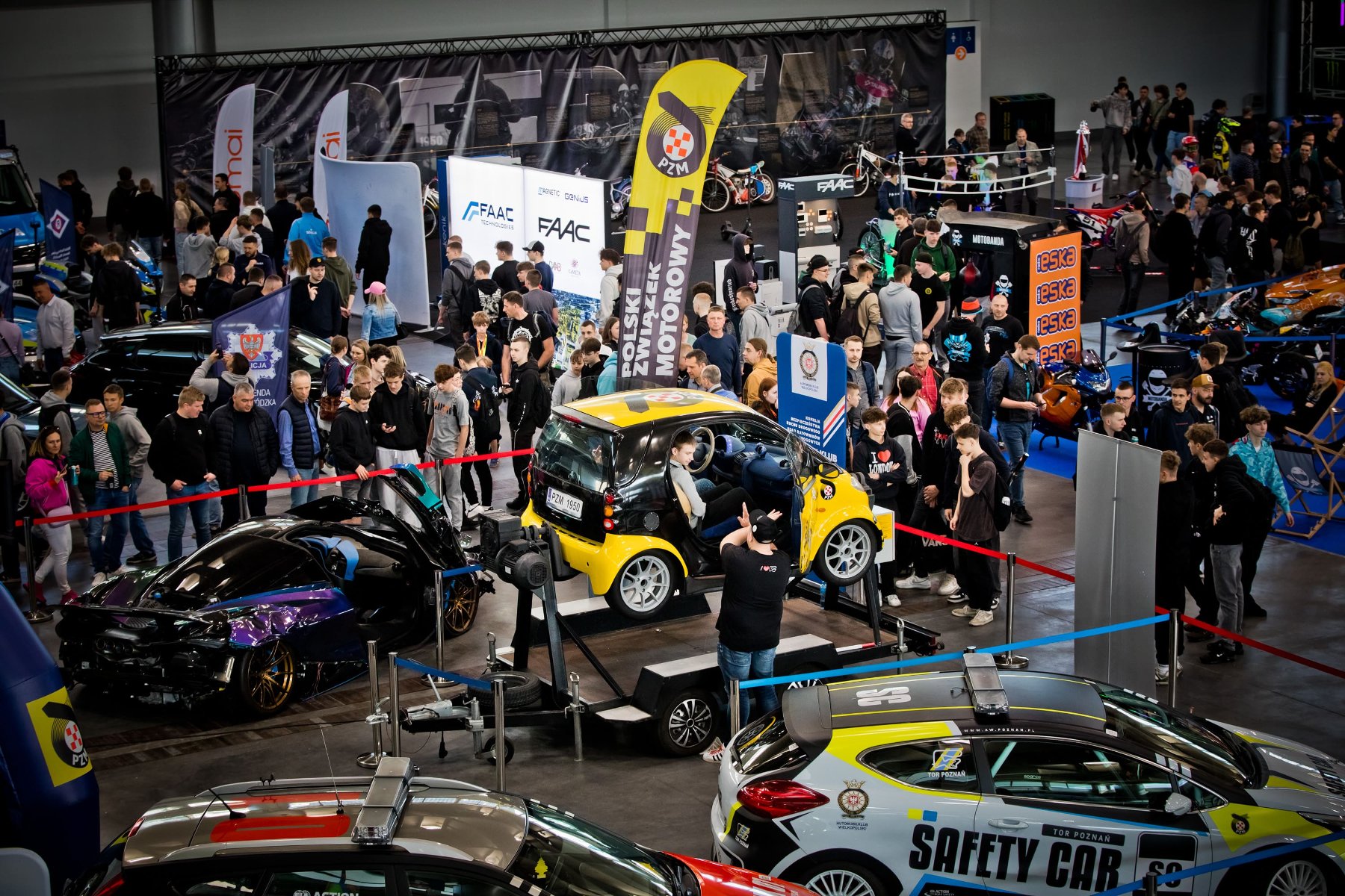 Zdjęcie przedstawia dynamiczną scenę z targów Poznań Motor Show. Na pierwszym planie, po lewej stronie, widoczny jest bok luksusowego, czerwonego samochodu sportowego o lśniącej karoserii. W centralnej części kadru znajduje się uśmiechnięta kobieta w eleganckim, ciemnym stroju, która trzyma mikrofon i prawdopodobnie przeprowadza wywiad lub relacjonuje wydarzenie