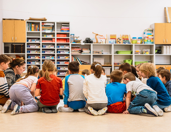 Zdjęcie przedstawia dzieci i nauczycielkę siedzących w kółku na dywanie w sali przedszkolnej. Dalej widać szafki z zabawkami.