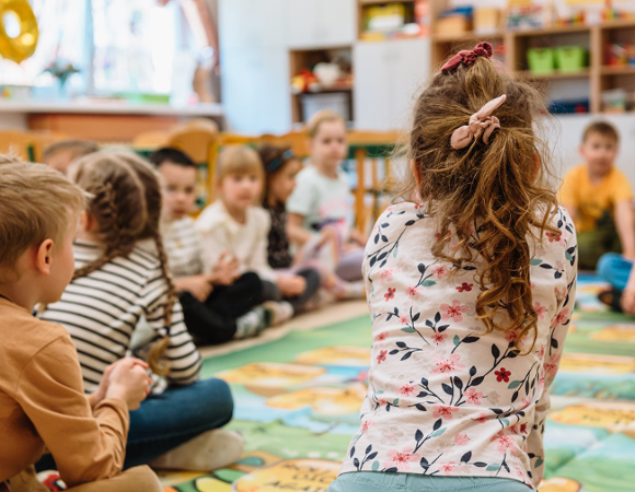 Zdjęcie przedstawia dzieci siedzące na dywanie w przedszkolnej sali.