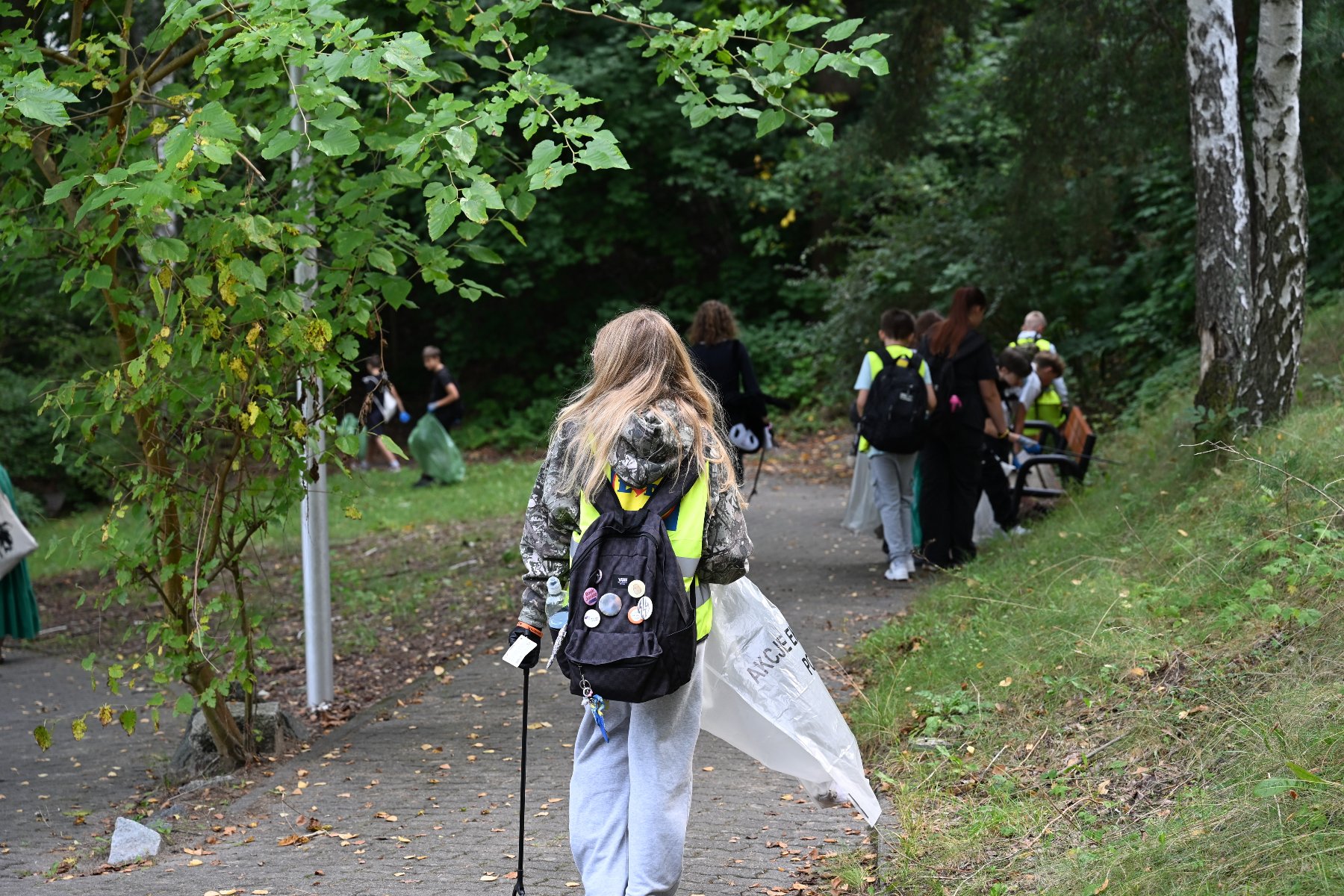 Zdjęcie przedstawia dziewczynę idącą z workiem na śmieci. Dalej widać innych młodych ludzi.