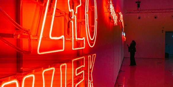 Zdjęcie przedstawia dziewczynę stojącą przy ścianie, na której umieszczonych jest kilka neonów.