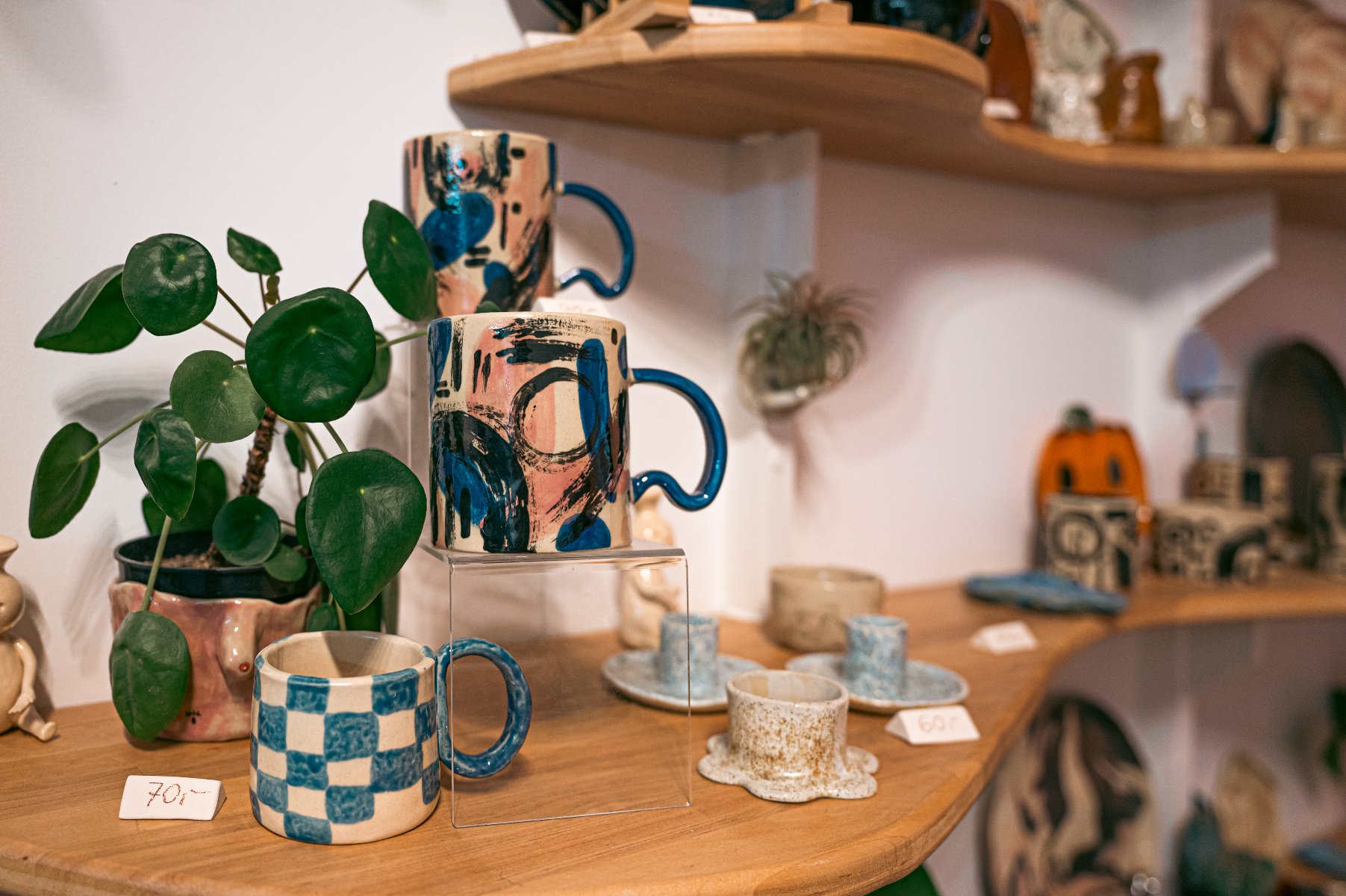 Zdjęcie przedstawia ekspozycję ręcznie robionej ceramiki na drewnianych, asymetrycznych półkach. Na pierwszym planie widoczne są kubki z charakterystycznymi niebieskimi uchwytami o nieregularnym kształcie, zdobione abstrakcyjnymi wzorami w odcieniach różu, beżu i błękitu. Obok nich stoi mniejszy kubek w niebiesko-białą szachownicę oraz doniczka z zieloną rośliną (Pilea).