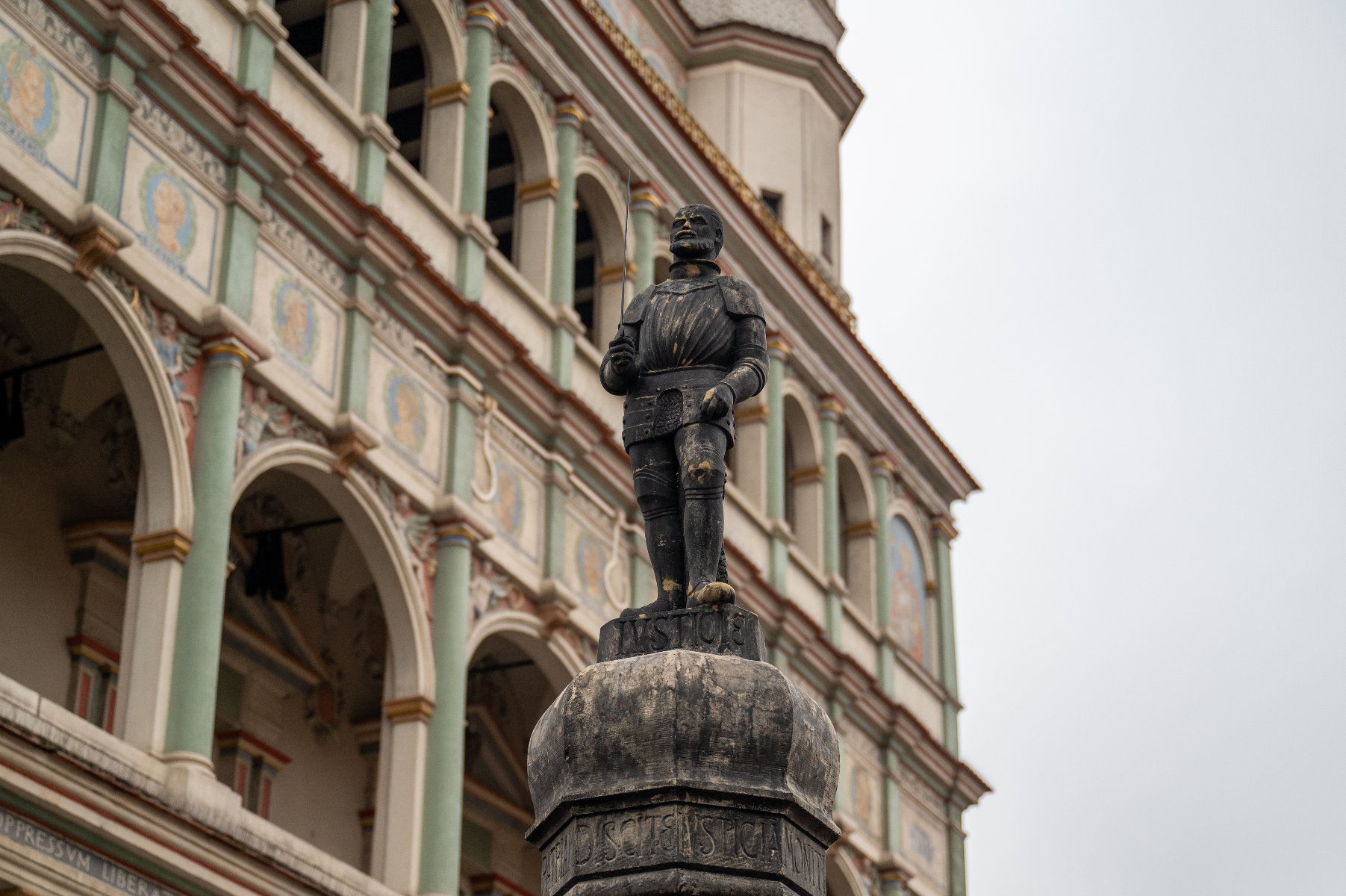 Zdjęcie przedstawia figurę kata z mieczem, wieńczącą poznański pręgierz na Starym Rynku. Rzeźba stoi na ozdobnej kolumnie z widocznym napisem "IUSTITIA". W tle wyłania się renesansowa fasada poznańskiego Ratusza z charakterystycznymi arkadami, kolumienkami oraz malowidłami. Niebo nad budowlą jest zachmurzone.