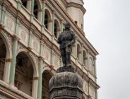 Zdjęcie przedstawia figurę kata z mieczem, wieńczącą poznański pręgierz na Starym Rynku. Rzeźba stoi na ozdobnej kolumnie z widocznym napisem "IUSTITIA". W tle wyłania się renesansowa fasada poznańskiego Ratusza z charakterystycznymi arkadami, kolumienkami oraz malowidłami. Niebo nad budowlą jest zachmurzone.