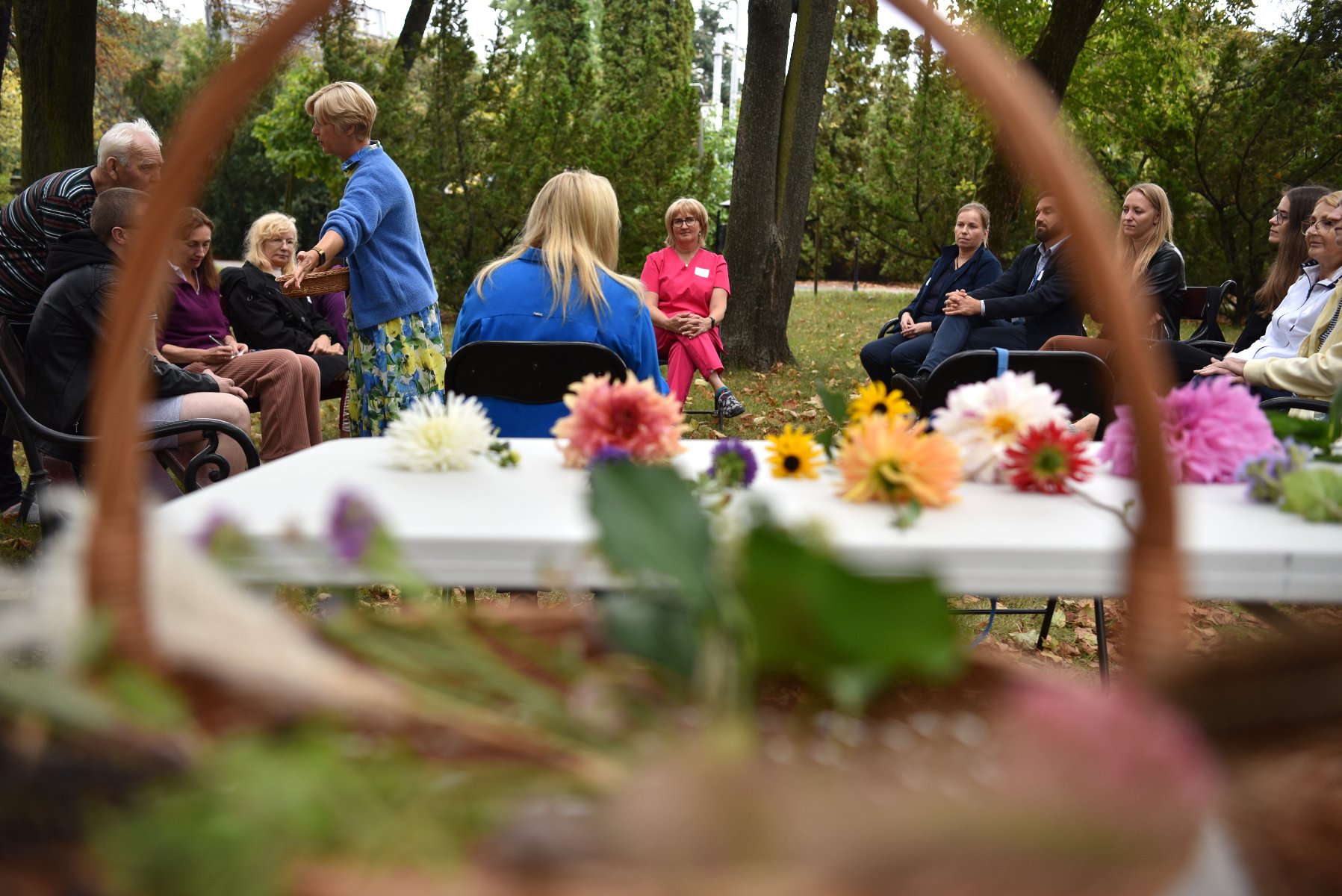 Zdjęcie przedstawia grupę ludzi siedzących na krzesłach wśród zieleni. Na pierwszym planie znajdują się kwiaty.