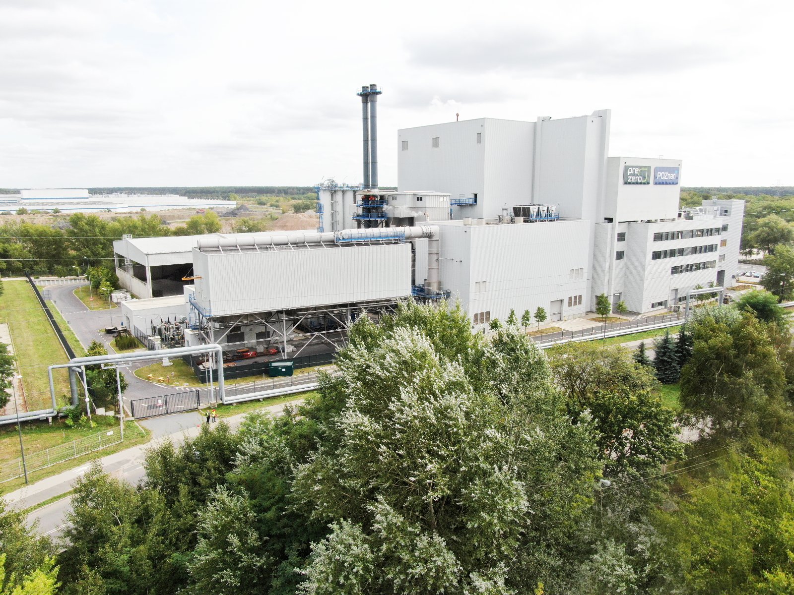 Zdjęcie przedstawia grupę ludzi w odblaskowych kamizelkach. Przed nimi widać budynek z dużym dymiącym kominem.