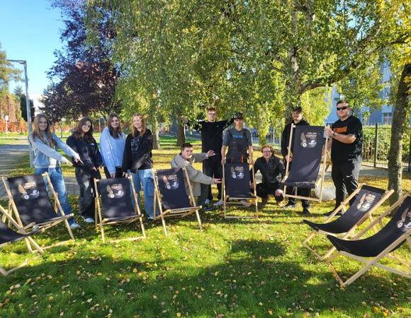 Zdjęcie przedstawia grupę młodych ludzi, którzy pokazują rękami na leżaki. Wokół nich jest zieleń.