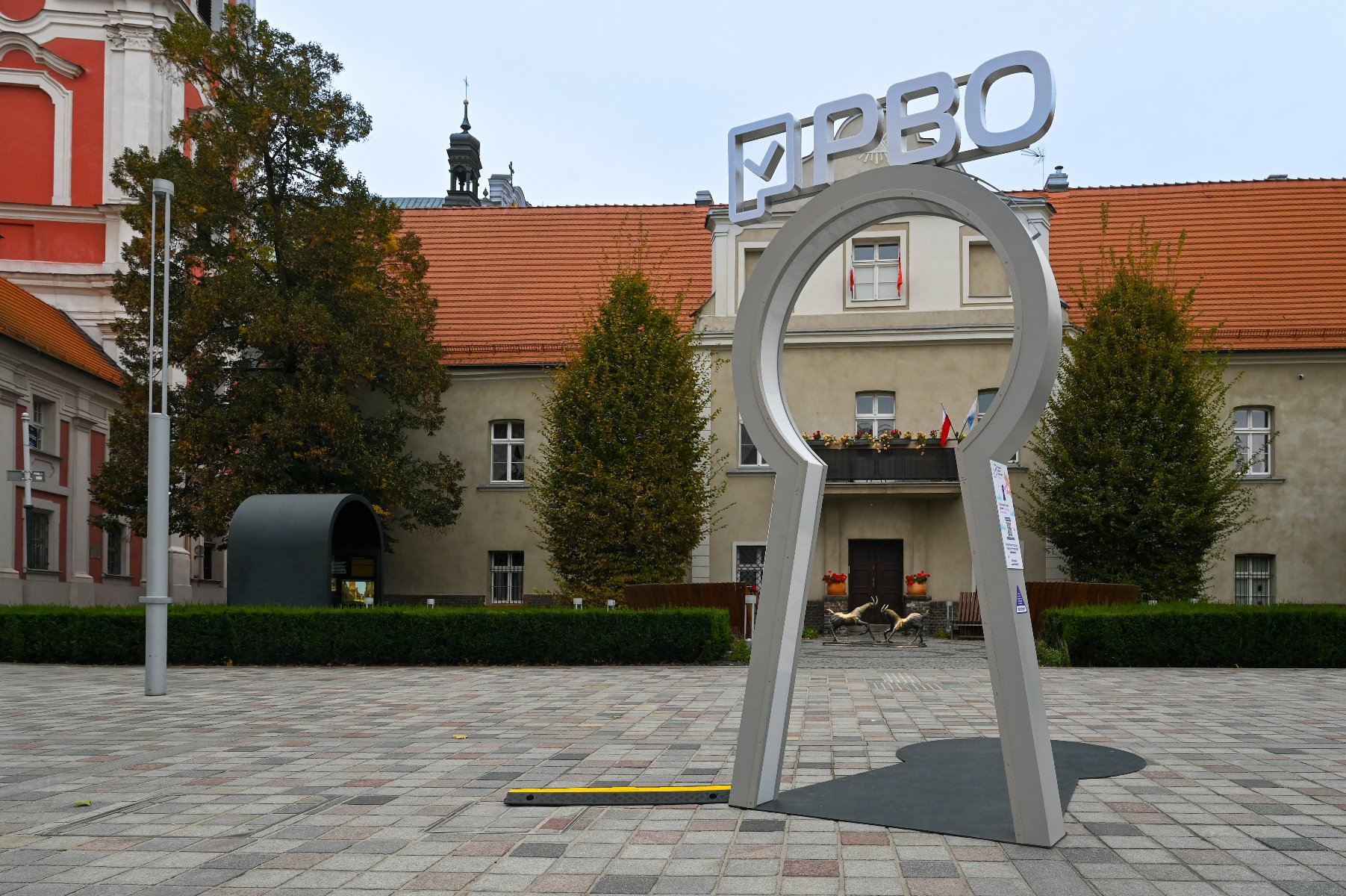 Zdjęcie przedstawia instalację w kształcie dużej dziurki od klucza, stojącej na placu. Na górze tej instalacji znajduje się napis PBO.