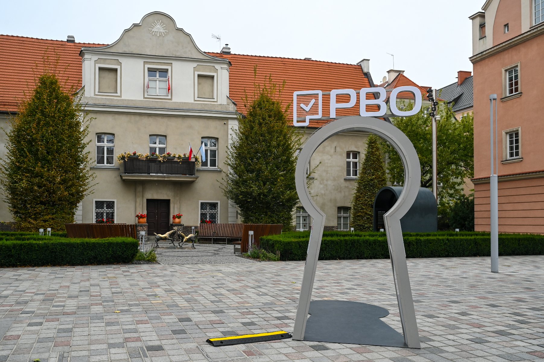 Zdjęcie przedstawia instalację w kształcie dużej dziurki od klucza, stojącej na placu. Na górze tej instalacji znajduje się napis PBO. Obok widać budynki oraz rzeźbę dwóch trykających się koziołków.