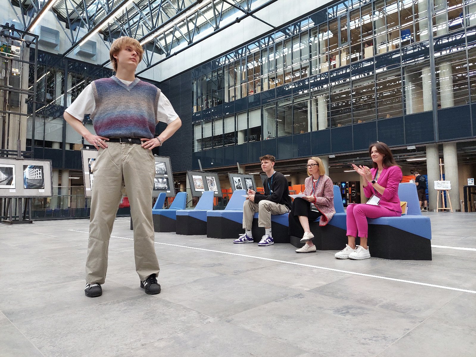 Zdjęcie przedstawia jednego z uczesników pokazu mody "Nici z planety", któy odbywa sie w holu Politechniki Poznańskiej. Mężczyzna stoi w lekkim rozkroku z rękoma opartymi na biodrach. W drugim planie widać trzy siedzące osoby. Jedna klaszcze.