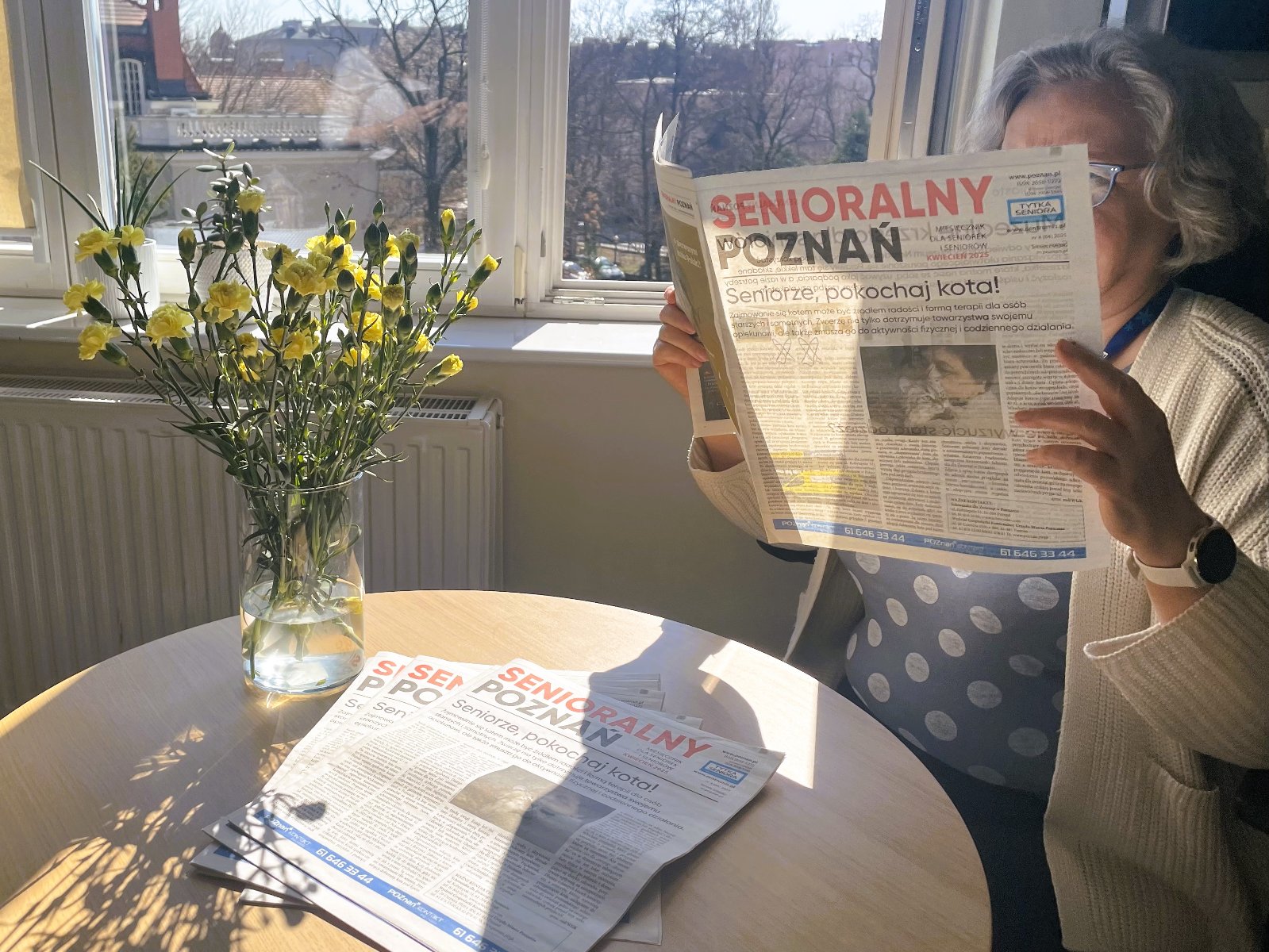 Zdjęcie przedstawia kobiete czytającą miesięcznik Senioralny Poznań w oświetlonej słońcem wiosennej scenerii