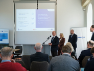 Zdjęcie przedstawia kobietę i dwóch mężczyzn stojących obok siebie. Jeden z mężczyzn stoi przed mikrofonem. Na krzesłach przed nimi siedzą ludzie.