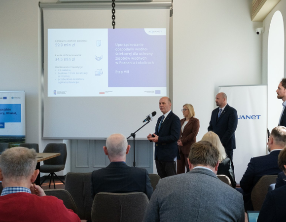 Zdjęcie przedstawia kobietę i dwóch mężczyzn stojących obok siebie. Jeden z mężczyzn stoi przed mikrofonem. Na krzesłach przed nimi siedzą ludzie.