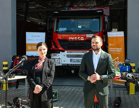 Zdjęcie przedstawia konferencję prasową lub oficjalną prezentację zorganizowaną przed garażem straży pożarnej. Na pierwszym planie stoi dwoje prelegentów: kobieta w czarnym żarcie z czerwoną apaszką oraz mężczyzna w ciemnozielonym garniturze. W tle, bezpośrednio za nimi, widoczny jest nowoczesny, czerwony wóz strażacki marki Iveco.