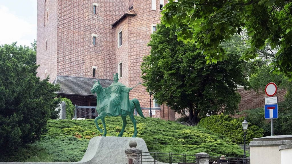 Zdjęcie przedstawia konny pomnik króla Przemysła II, który znajduje się na Górze Przemysła w Poznaniu. Zielona rzeźba stoi na postumencie na tle gęstych krzewów i drzew. W tle widoczny jest fragment zamku z czerwonej cegły (Zamek Królewski w Poznaniu) oraz znak drogowy informujący o ślepej uliczce.