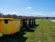 Zdjęcie przedstawia kontenery na odpady, które stoją na terenie zielonym. W oddali widać most.
