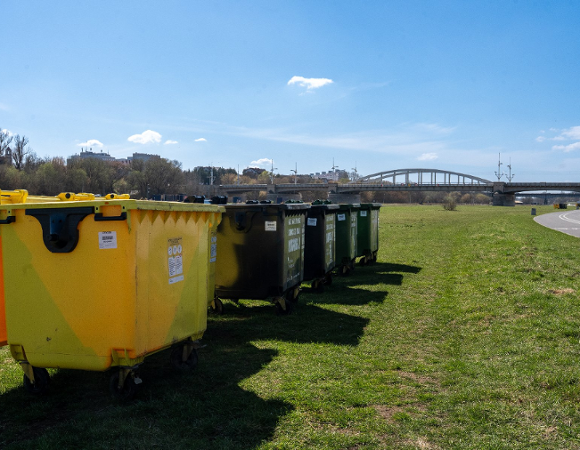 Zdjęcie przedstawia kontenery na odpady, które stoją na terenie zielonym. W oddali widać most.
