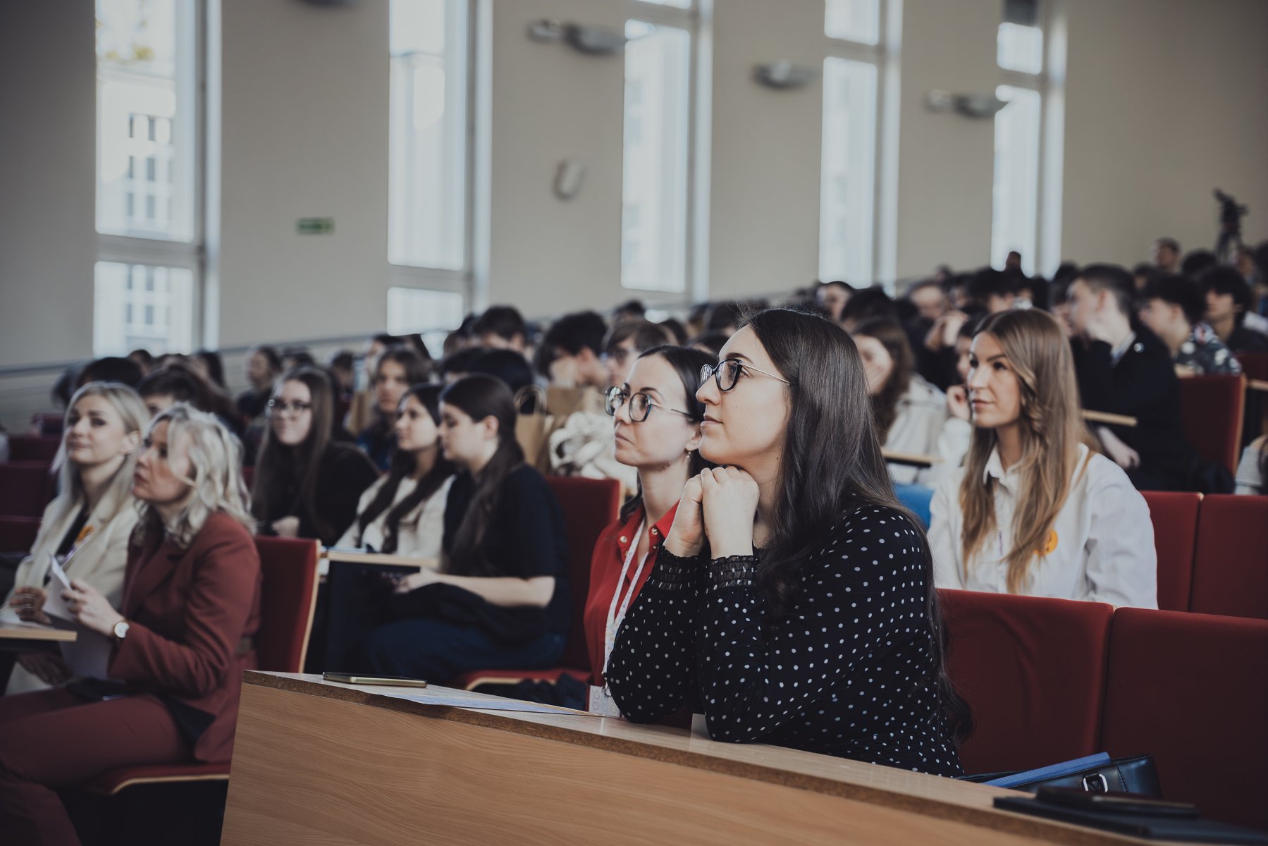 Zdjęcie przedstawia liczną publiczność siedzącą w rzędach auli wykładowej lub sali konferencyjnej. Na pierwszym planie widać kobietę w ciemnej bluzce w białe kropki, która z uwagą patrzy przed siebie, opierając dłonie na blacie.