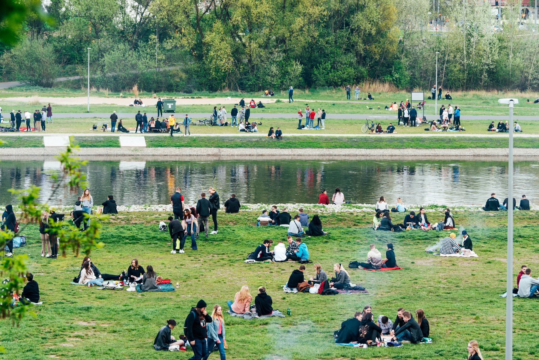 Zdjęcie przedstawia liczne grupy osób wypoczywających na trawiastym brzegu rzeki Warty w Poznaniu. Ludzie siedzą na kocach, piknikują i rozmawiają, ciesząc się słoneczną pogodą. W tle widać koryto rzeki oraz przeciwległy brzeg z gęstą roślinnością i drzewami.