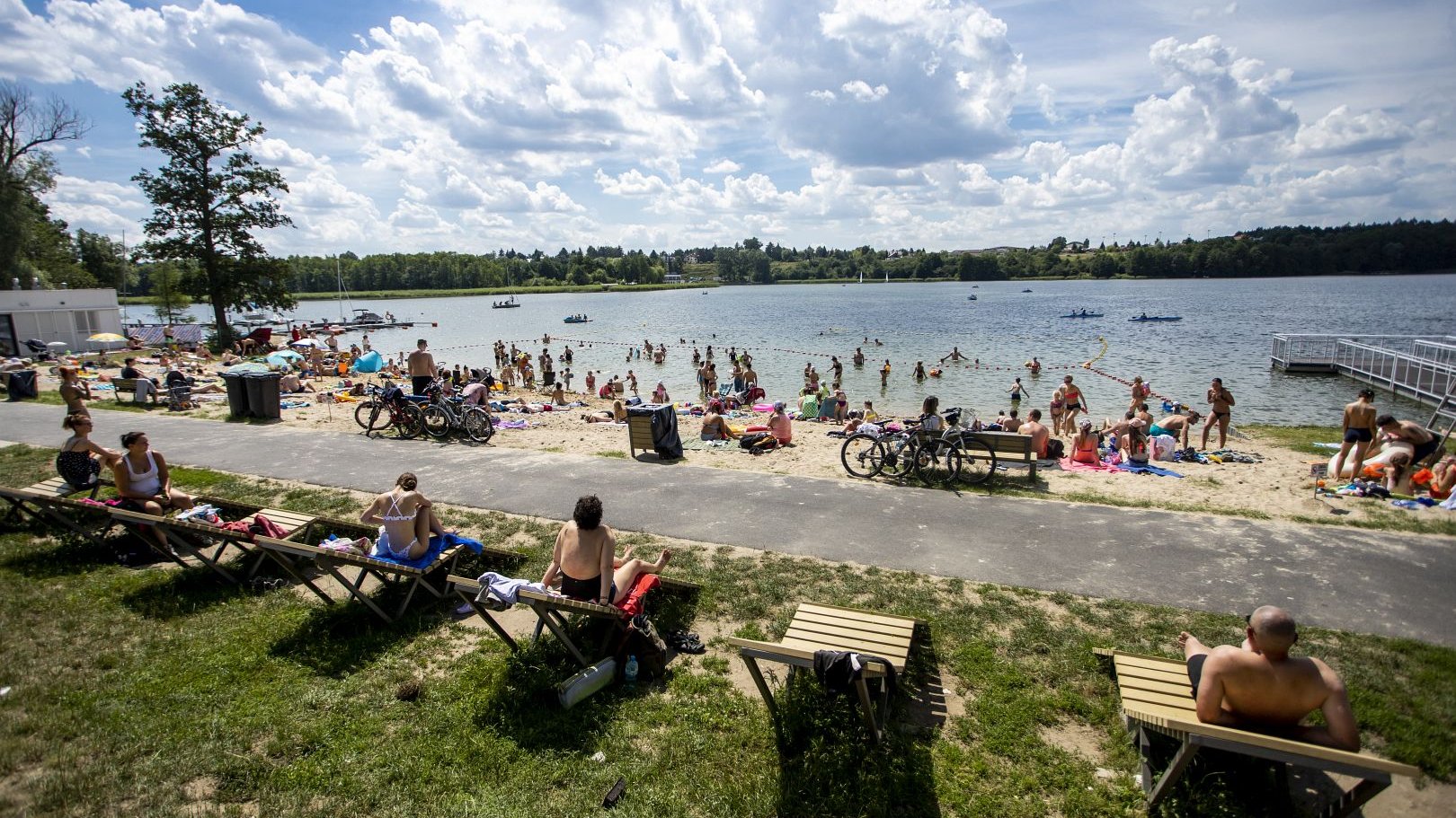Zdjęcie przedstawia ludzi leżacych na leżakach i na kocach na plaży. Dalej widać jezioro i pływających w nim ludzi.