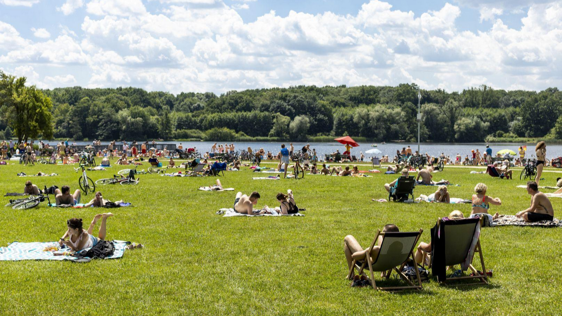 Zdjęcie przedstawia ludzi odpoczywających na trawiastej plaży. W tle widać jezioro.