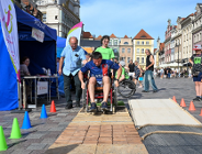 Zdjęcie przedstawia mężczyznę jadącego na wózku po torze. Zdjęcie zrobiono na Starym Rynku. W tle widać namioty, kamienicę i innych ludzi.