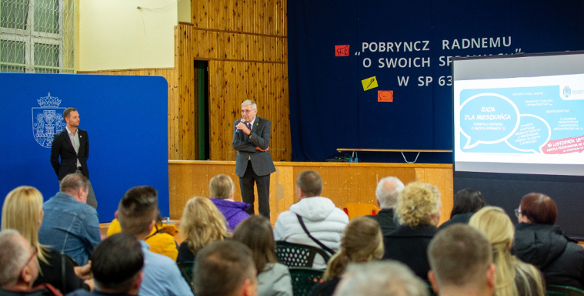 Zdjęcie przedstawia mężczyznę przy mikrofonie i tłum ludzi siedzących na krzesłach przed nim.