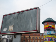 Zdjęcie przedstawia miejskie nośniki reklamowe na tle pochmurnego nieba i fragmentów budynków. Po lewej stronie znajduje się duży, pusty billboard reklamowy o metalowej, pionowo żłobionej strukturze z czerwoną ramą, zamontowany na grubym słupie. W jego dolnym lewym rogu naklejona jest mała tabliczka informacyjna. Po prawej stronie stoi okrągły słup ogłoszeniowy z zielonym daszkiem, oklejony kolorowymi plakatami.