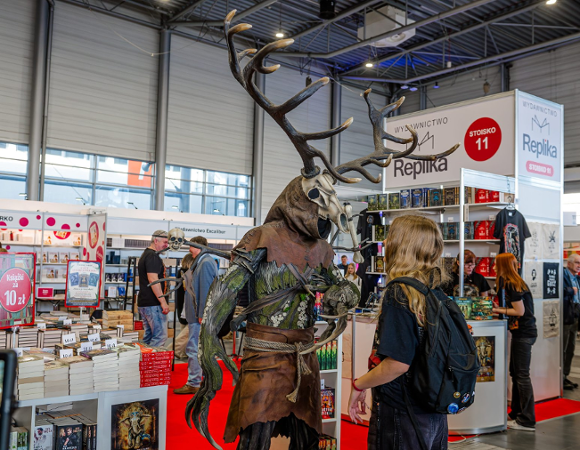 Zdjęcie przedstawia młodą osobę stojącą na przeciwko człowieka przebranego za Leszego. W tle widać stoiska z książkami i ludzi.