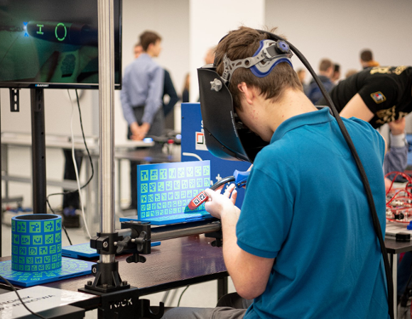 Zdjęcie przedstawia młodego mężczyznę w niebieskiej koszulce polo, korzystającego z symulatora spawania w rozszerzonej rzeczywistości. Uczeń ma na głowie przyłbicę i operuje uchwytem spawalniczym nad elementem oznaczonym zielonymi kodami. W tle widać monitor z podglądem pracy oraz stanowisko opisane jako "Technik Spawalnictwa", co wskazuje na charakter edukacyjny wydarzenia.