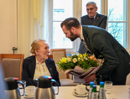 Zdjęcie przedstawia młodego mężczyznę wręczającego kwiaty starszej kobiecie siedzącej za stołem.