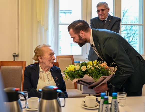 Zdjęcie przedstawia młodego mężczyznę wręczającego kwiaty starszej kobiecie siedzącej za stołem.