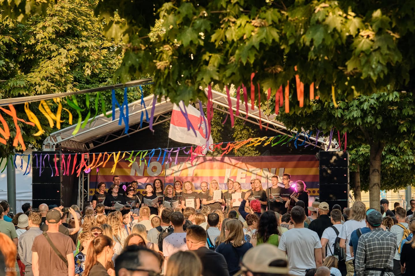Zdjęcie przedstawia młodych artystów na scenie. Stoją tyłem do aparatu, a przodem do widowni, trzymają w ręce książki z nutami. Pod sceną stoi tłum ludzi. Scena znajduje się na zewnątrz.