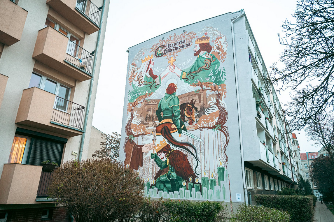 Zdjęcie przedstawia mural na budynku. Mural podzielony jest na 3 częśći. Na górze widnieje napis "Kronika Galla Anonima" oraz rysunek przedstawiający mężczyznę i kobietę w koronach, trzymających wspólnie krzyż. Na środku widać mężczyznę na koniu, siedzącego tyłem. Na dole postać mężczyzny, który się pochyla.
