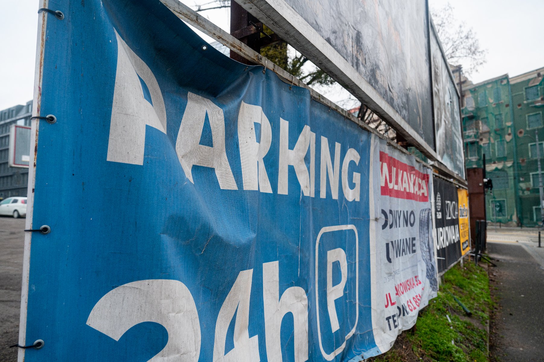 Zdjęcie przedstawia niebieski baner reklamowy z białym napisem "PARKING 24h", zamocowany na metalowym ogrodzeniu. Baner jest lekko pognieciony i przymocowany za pomocą czarnych opasek zaciskowych. W tle, za niebieską płachtą, widoczne są kolejne tablice reklamowe oraz fragment ulicy z chodnikiem i budynkiem w oddali. Perspektywa zdjęcia jest ujęta pod kątem, co podkreśla długość ogrodzenia oddzielającego parking od reszty terenu.