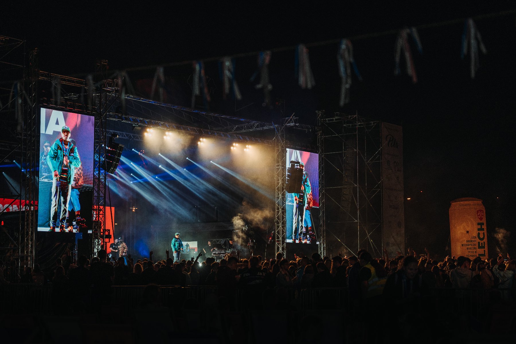 Zdjęcie przedstawia nocny koncert plenerowy na dużej scenie z profesjonalnym nagłośnieniem i oświetleniem. Na środku występuje artysta w turkusowej kurtce, którego wizerunek jest jednocześnie wyświetlany na dwóch wielkich telebimach po bokach sceny. Przed estradą zgromadził się liczny tłum fanów, a dynamiczne snopy światła i dym sceniczny tworzą widowiskową oprawę muzycznego wydarzenia.
