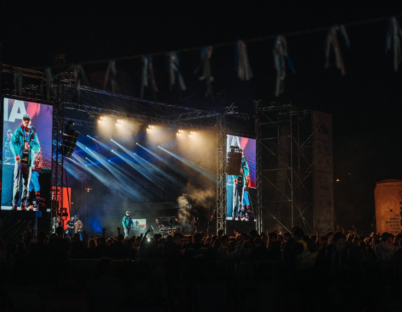 Zdjęcie przedstawia nocny koncert plenerowy na dużej scenie z profesjonalnym nagłośnieniem i oświetleniem. Na środku występuje artysta w turkusowej kurtce, którego wizerunek jest jednocześnie wyświetlany na dwóch wielkich telebimach po bokach sceny. Przed estradą zgromadził się liczny tłum fanów, a dynamiczne snopy światła i dym sceniczny tworzą widowiskową oprawę muzycznego wydarzenia.