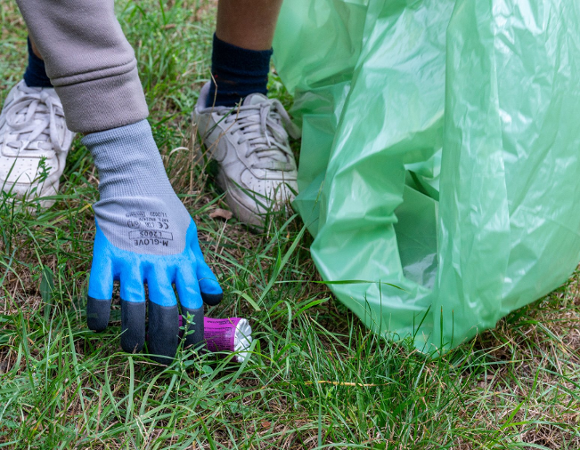 Zdjęcie przedstawia nogi i ręce osoby podnoszącej z ziemi śmieć, by wrzucić do do trzymanego w ręku worka.