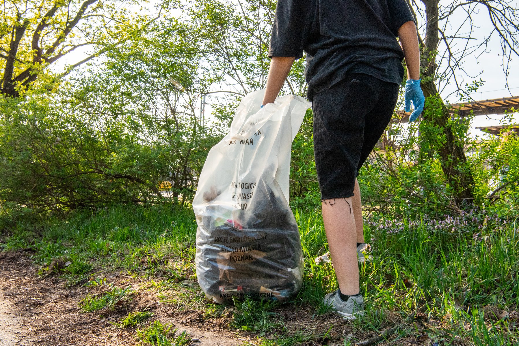Zdjęcie przedstawia nogi osoby sprzatającej, która w ręce trzyma worek ze śmieciami. Obok widać zieleń, drzewa i krzewy.