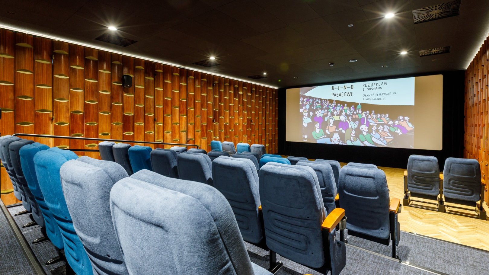 Zdjęcie przedstawia nowoczesną, kameralną salę Kina Pałacowego w Poznaniu, w której znajdują się rzędy wygodnych, niebieskich foteli skierowanych w stronę dużego ekranu projekcyjnego wyświetlającego ilustrowaną grafikę z napisem "K-I-N-O PAŁACOWE - BEZ REKLAM i popcornu".
